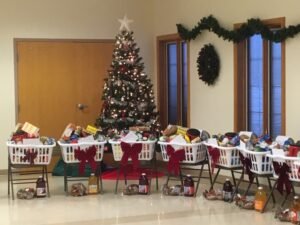 Clothes baskets sitting on metal folding chairs in a circle containing food items. A bow adorns the outside and a Christmas tree stands behind them.