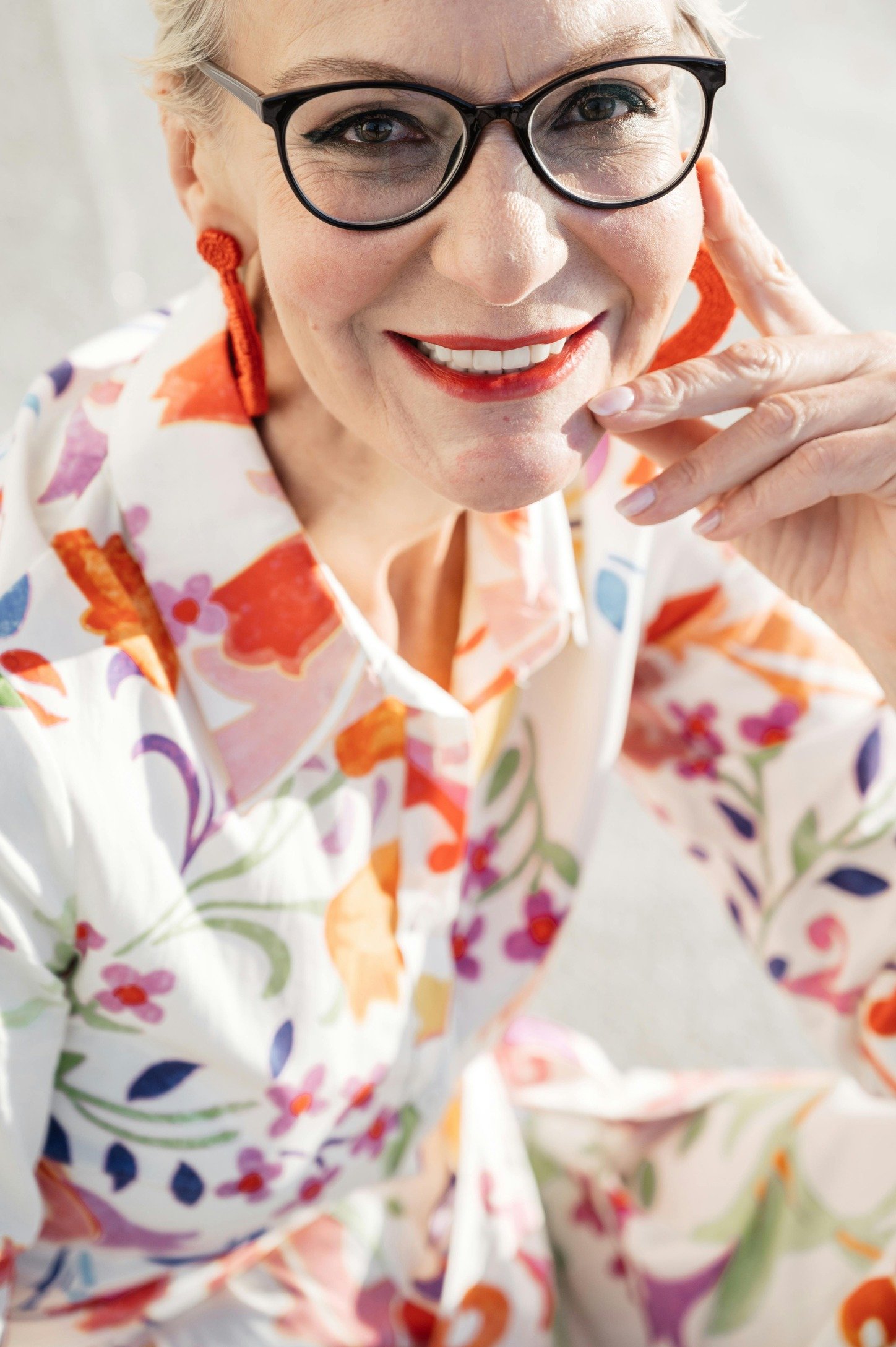 A senior woman wearing bright floral blouse, smiling with glasses.