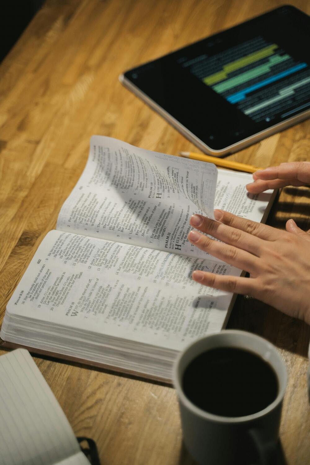 An open book with highlighted text, a person's hand turning a page, a cup of coffee, a tablet with notes, and a notebook on a wooden surface.