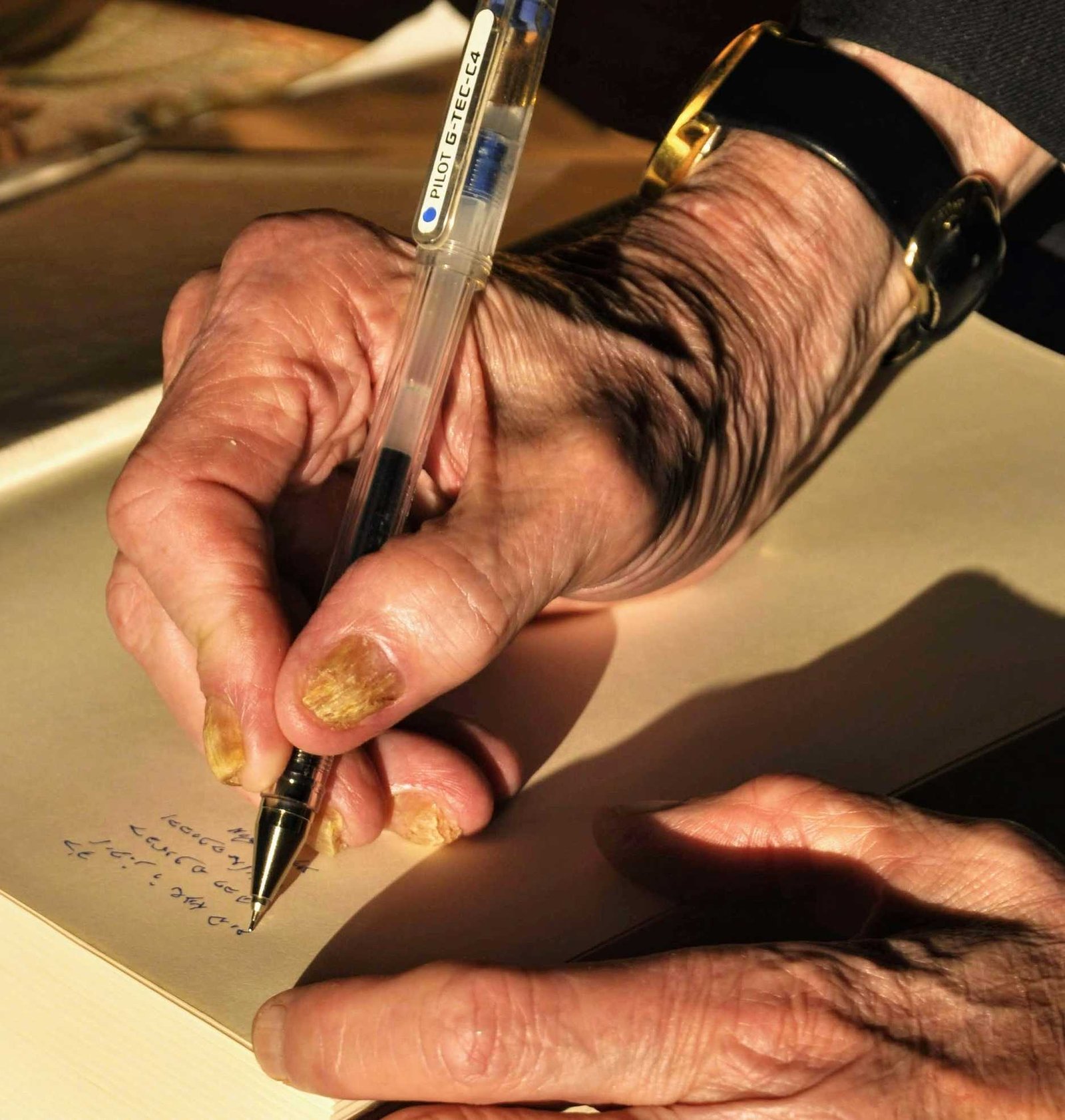 Close-up of an elderly person’s hands writing with a transparent ballpoint pen on beige paper.