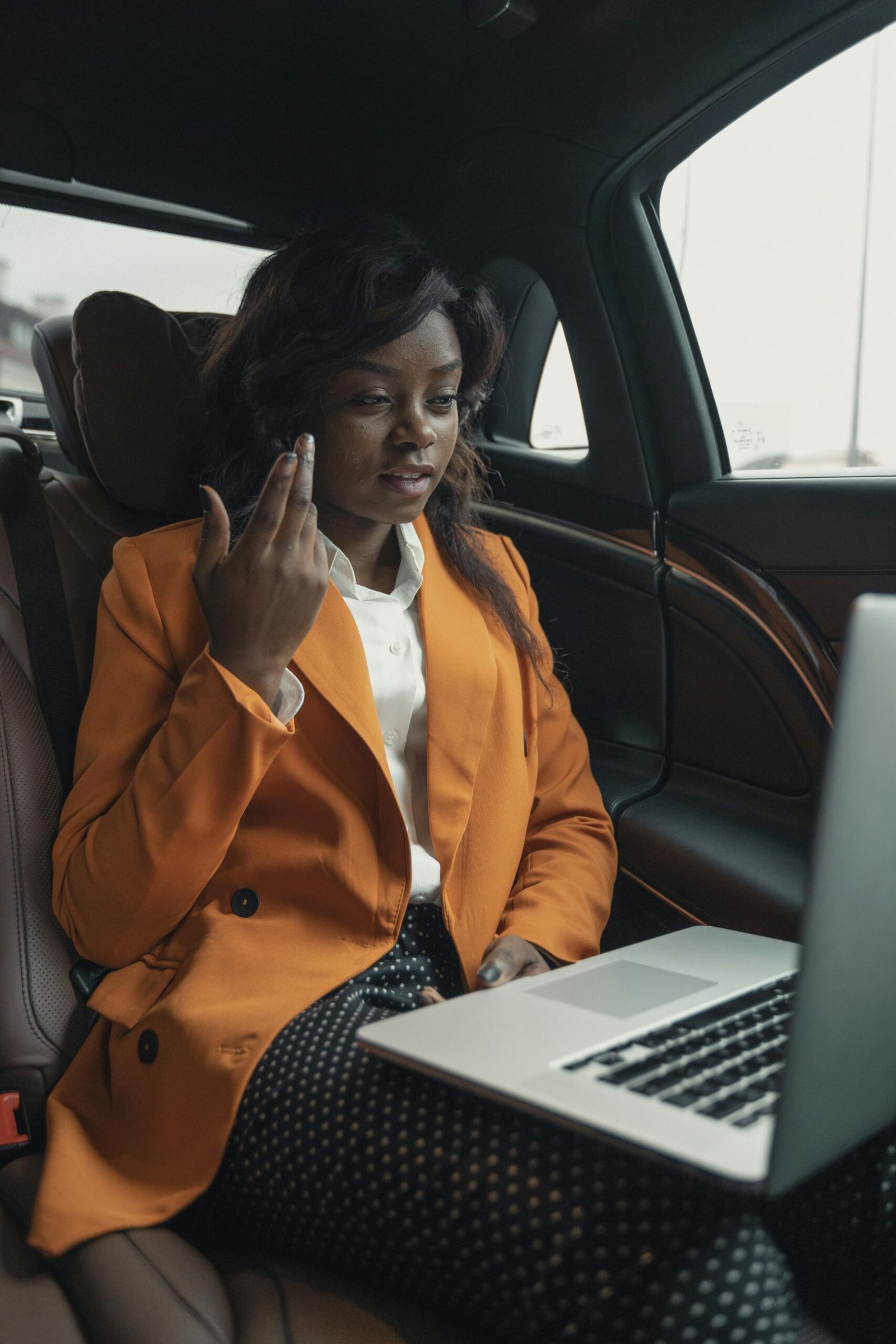 Woman in orange blazer sitting in a car back seat using a laptop.