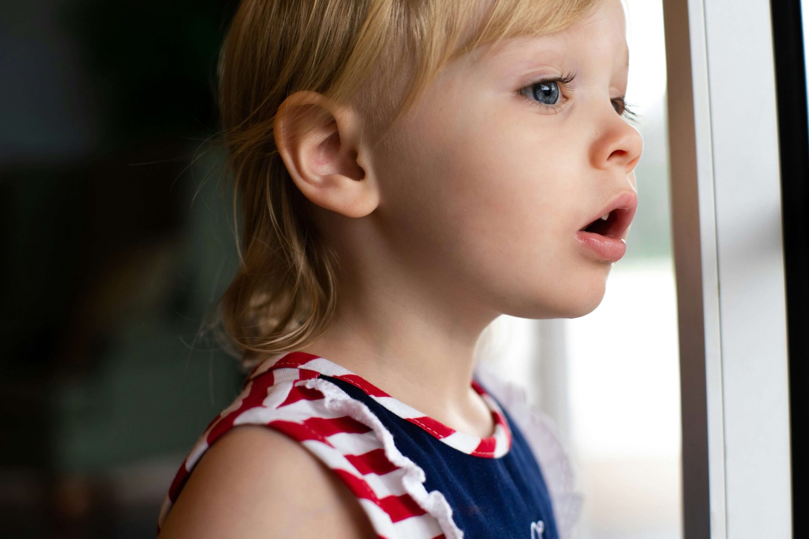 A young child with light hair looks out of a window with curiosity.